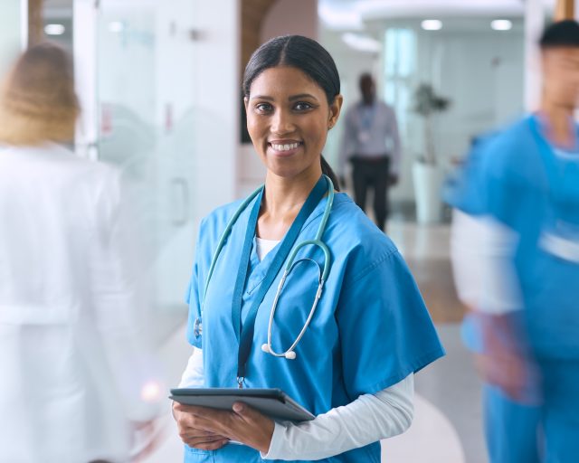 Portrait Of Female Doctor Or Nurse In Scrubs In Hospital Corridor With Colleagues Behind